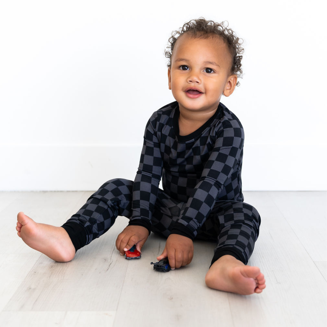 Toddler wearing black checkered soft bamboo two-piece pajama set sitting on floor playing with toy cars