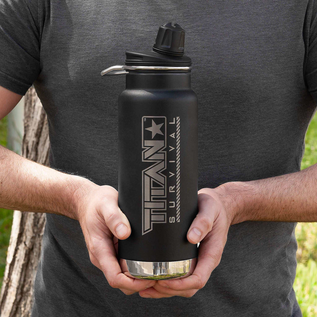 32 Oz wide mouth water bottle by TITAN Survival, vacuum insulated, black, double-walled, laser-etched logo, held by person outdoors.