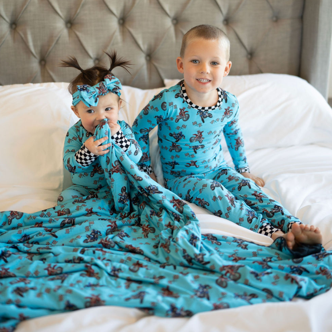 Two children wearing matching blue pajamas with a bamboo headband on the baby girl sitting on a bed
