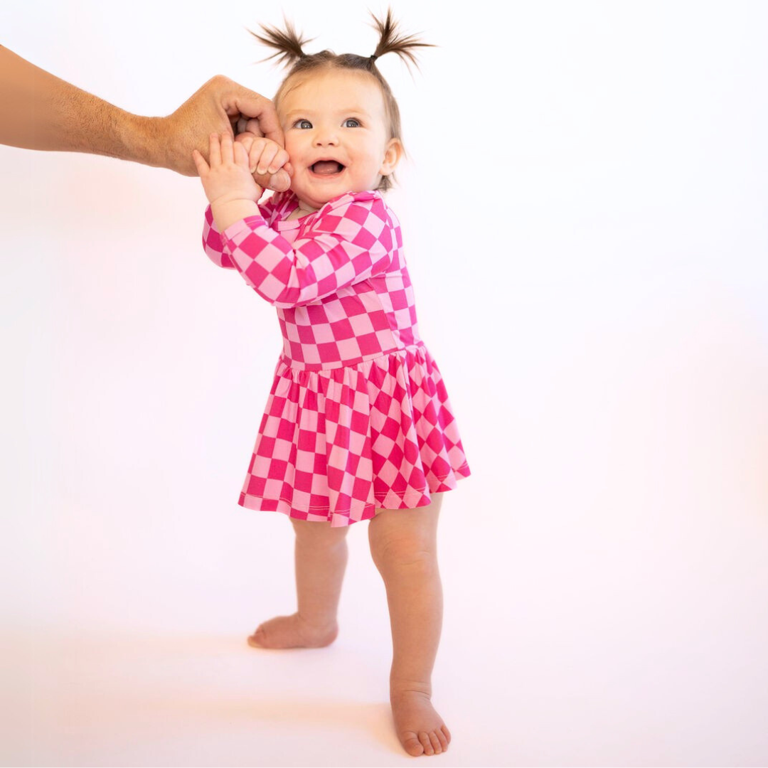 Baby wearing pink checkered ruffle dress long sleeve bodysuit with snap closures standing and smiling