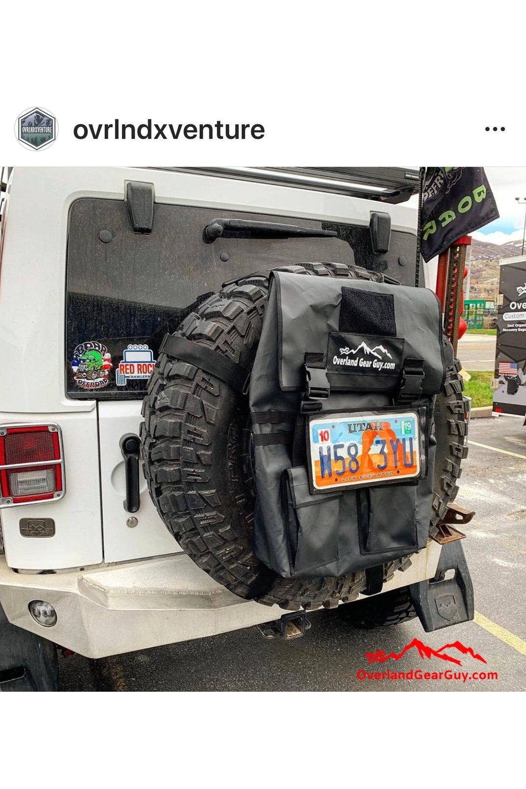 Black MOLLE license plate holder pouch attached to a spare tire on a white Jeep, featuring clear front sleeve and side Velcro closure.