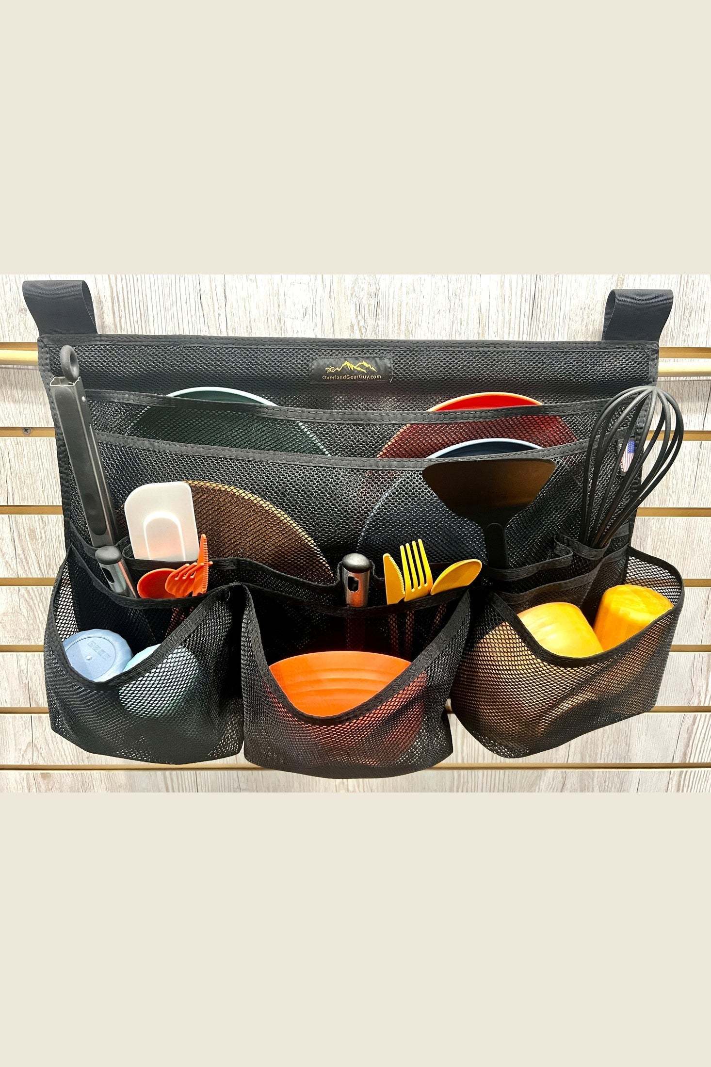 Black mesh dish drainer organizer hanging on wooden wall with plates, utensils, and kitchen tools inside compartments