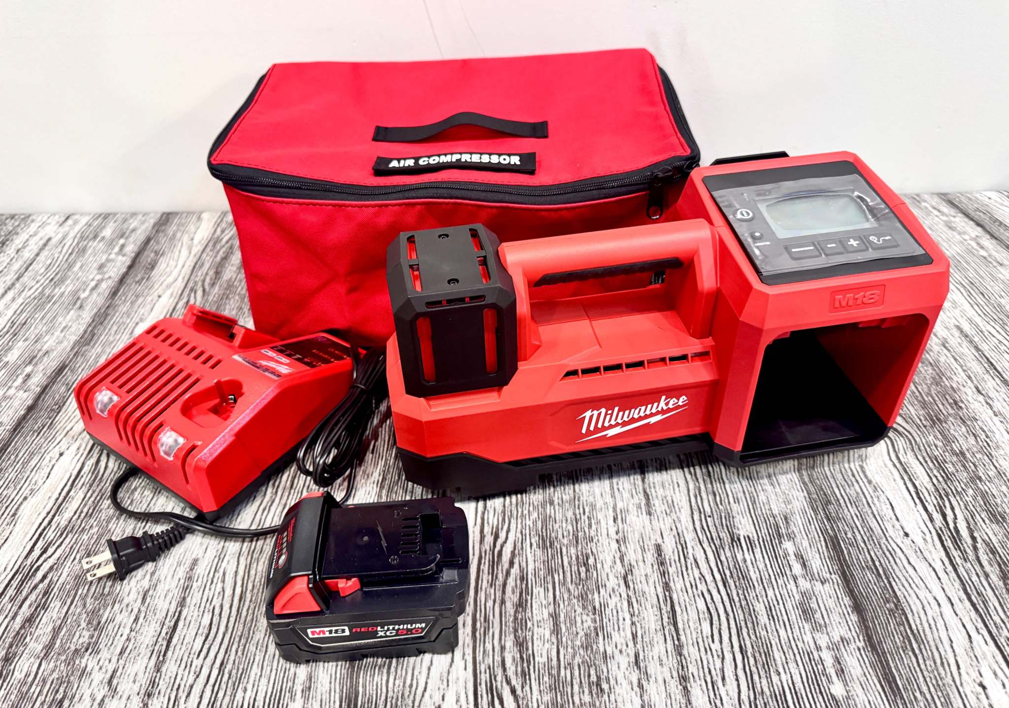 Milwaukee M18 Inflator with red AIR COMPRESSOR carrying bag, charger, and battery on wooden surface