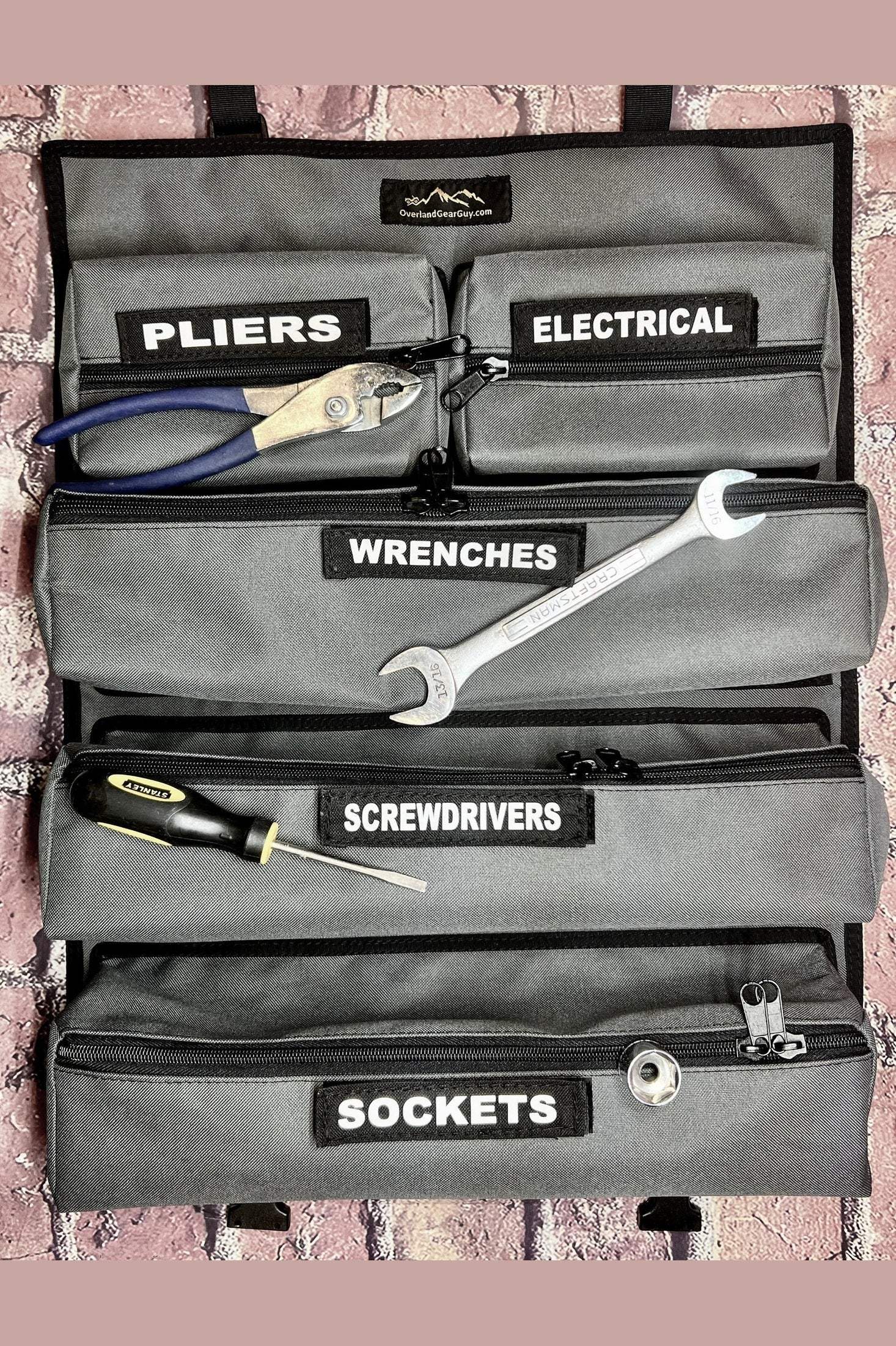 OGG Tool Roll with detachable pockets labeled for pliers, electrical, wrenches, screwdrivers, and sockets containing various tools organized on a brick background