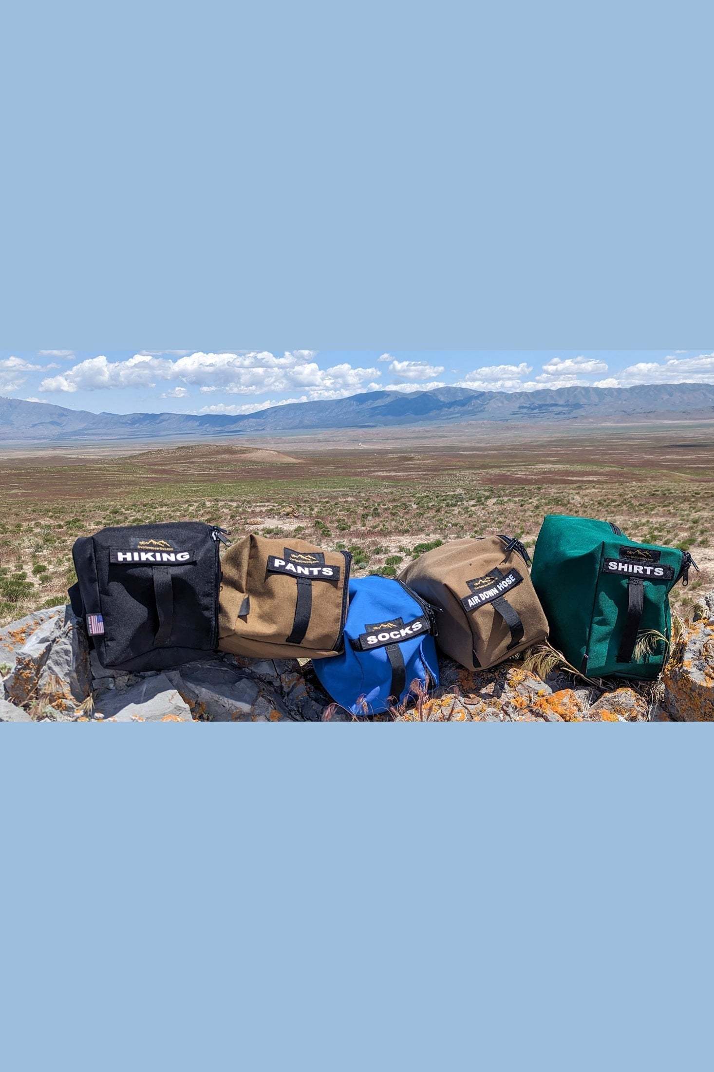 OGG Travel Bags in various colors and sizes labeled for hiking gear, pants, socks, tools, and shirts displayed outdoors on rocky terrain with a scenic mountain landscape background