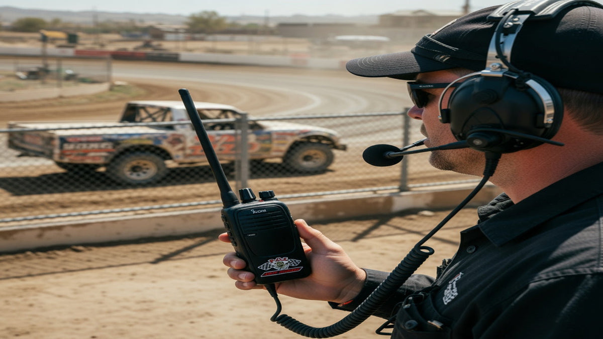 Short-course spotter in a crew-chief headset with a handheld radio beside a dirt-track catch fence