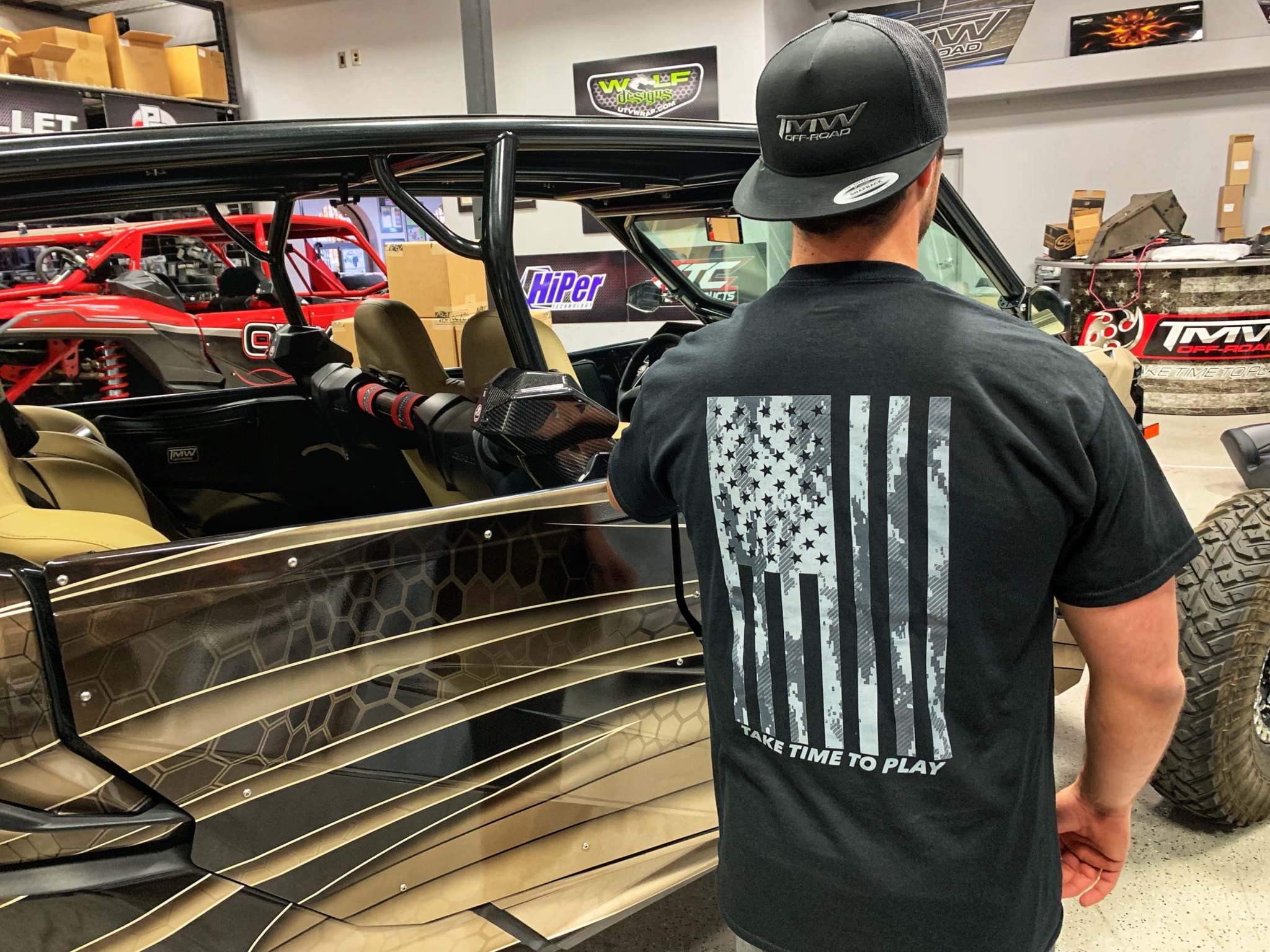Man wearing black TMW American Flag Camo tee with camo-style US flag and "Take Time To Play" text on back in workshop background